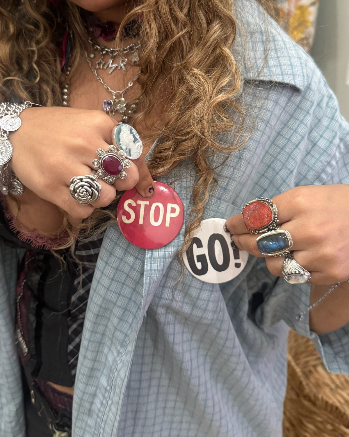 A close-up lifestyle shot of a woman with curly hair wearing silver-toned rings and pinning two protest-style buttons to her light blue plaid shirt. One button is pink with 'STOP' in white, and the other is white with 'GO!' in black.