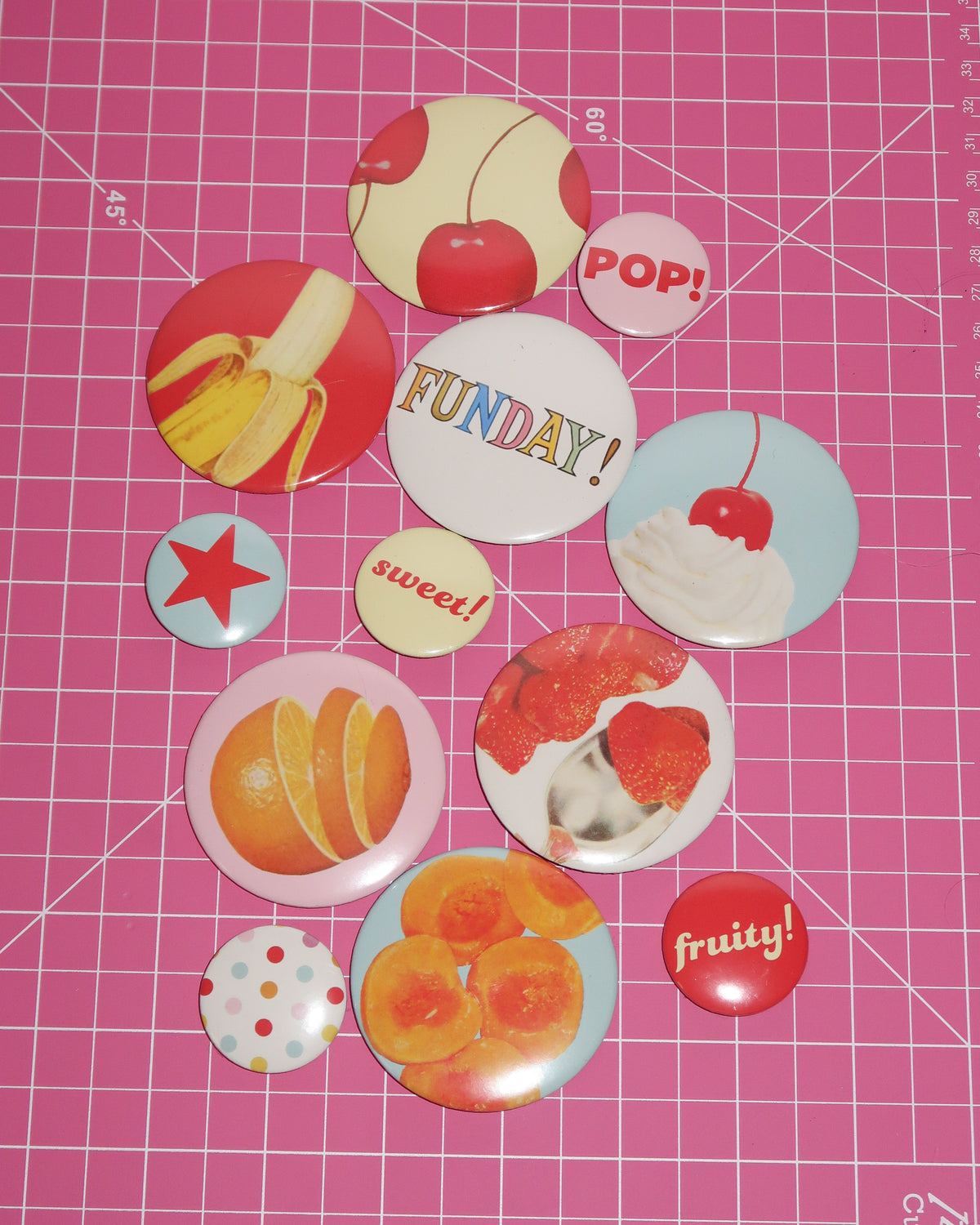 A set of colorful fruit-themed button badges arranged on a pink cutting mat with grid lines. The badges feature illustrations of cherries, bananas, strawberries, oranges, peaches, and text like 'FUNDAY!', 'sweet!', 'POP!', and 'fruity!'.