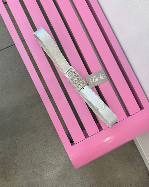A white shoulder bag with a rhinestone-embellished rectangular clasp and a silver-tone 'Funko' tag, displayed on a pink slatted bench.