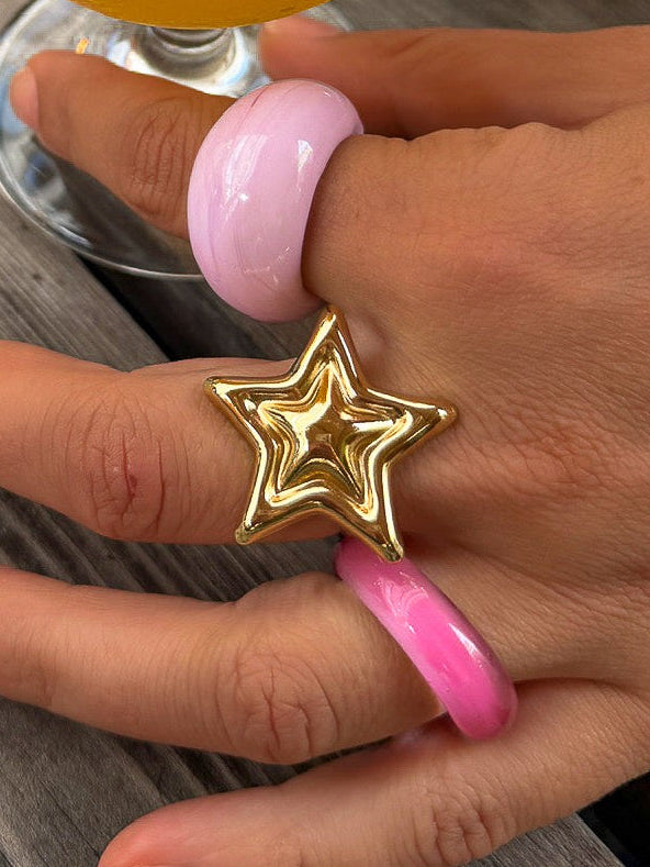 A hand wearing pink and gold rings rests on a weathered gray wooden table, holding a glass of frothy orange beverage. A gold clutch bag is visible in the background.