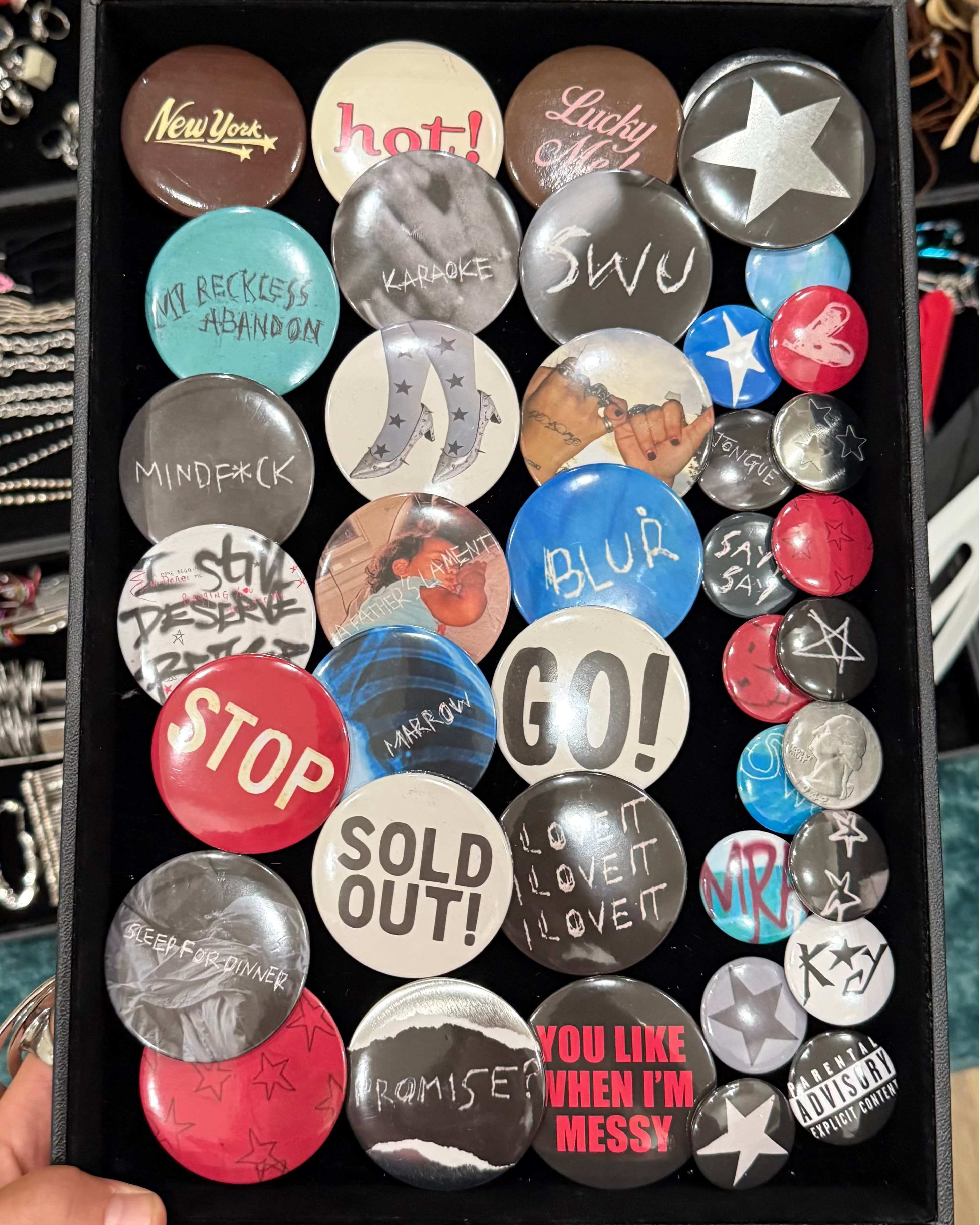 A hand holds a black tray filled with multiple round pin-back buttons featuring various designs, slogans, and graphics, with jewelry bins visible in the background.