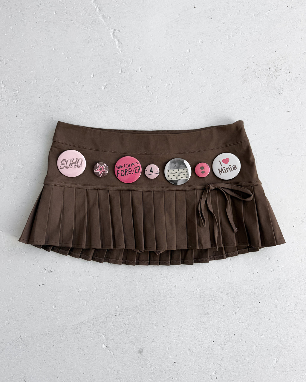 A brown pleated mini skirt with a tie waistband decorated with various colorful round and star-shaped pins, laid flat on a textured light gray background.