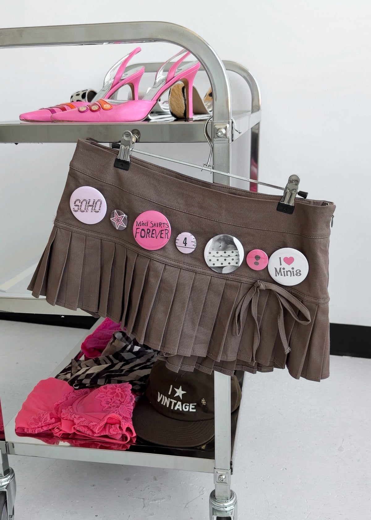 A brown pleated mini skirt with tie details and decorative buttons, hanging on a metal cart alongside pink high-heeled shoes and folded clothing.