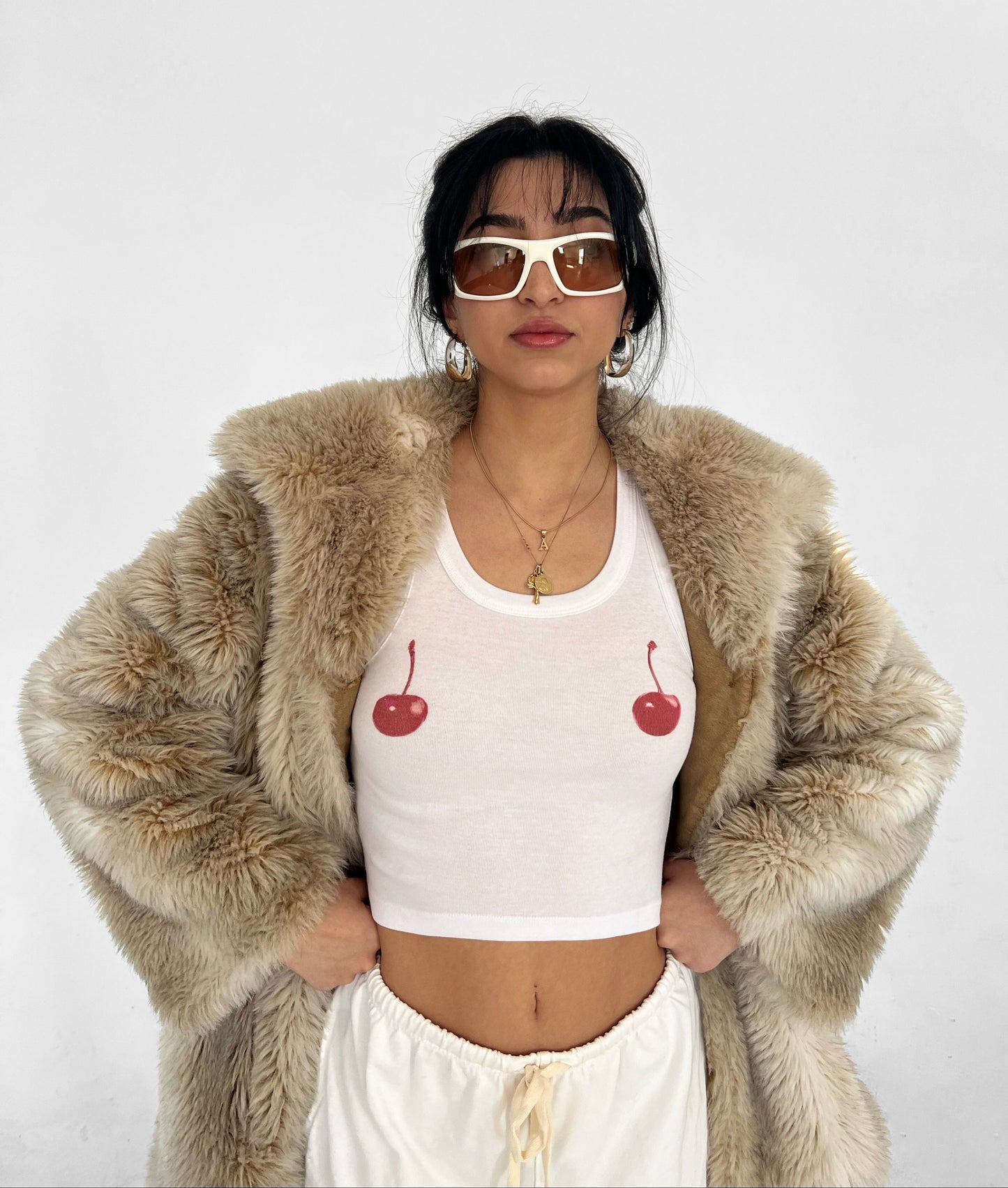 A woman wearing a white tank top with cherry graphics on the front, paired with a beige faux fur coat, sunglasses, and white pants.